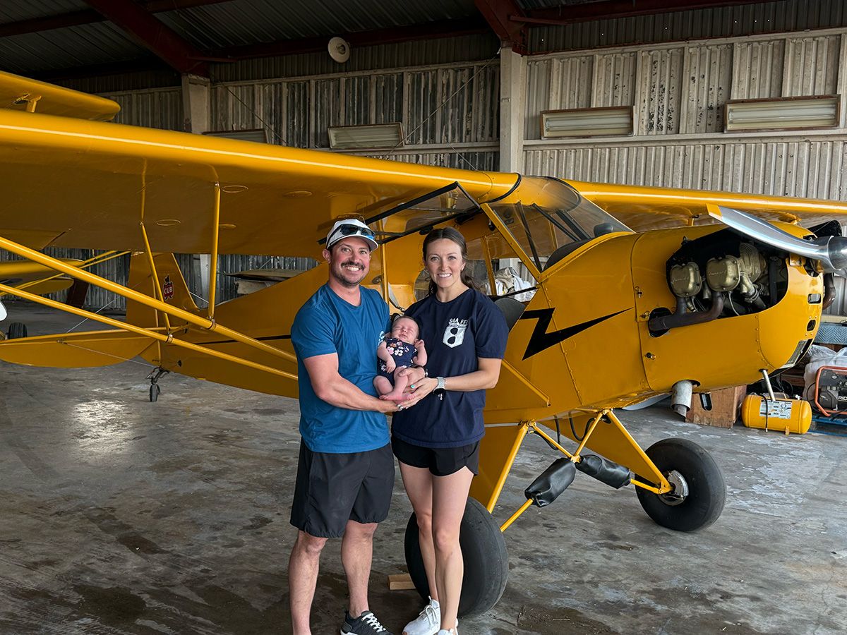 john cournoyer in blue shirt and brown felt fedora in front of a red biplane with yellow wings
